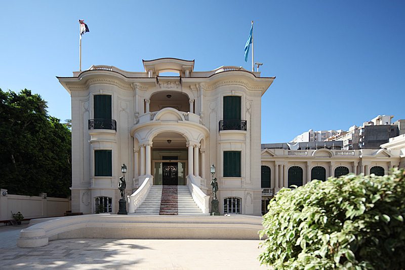 الواجهة البيضاء للجناح الغربي من قصر فاطمة الزهراء، بشرفاته ودرج المدخل وتمثالي حاملي المصابيح، تحت سماء زرقاء